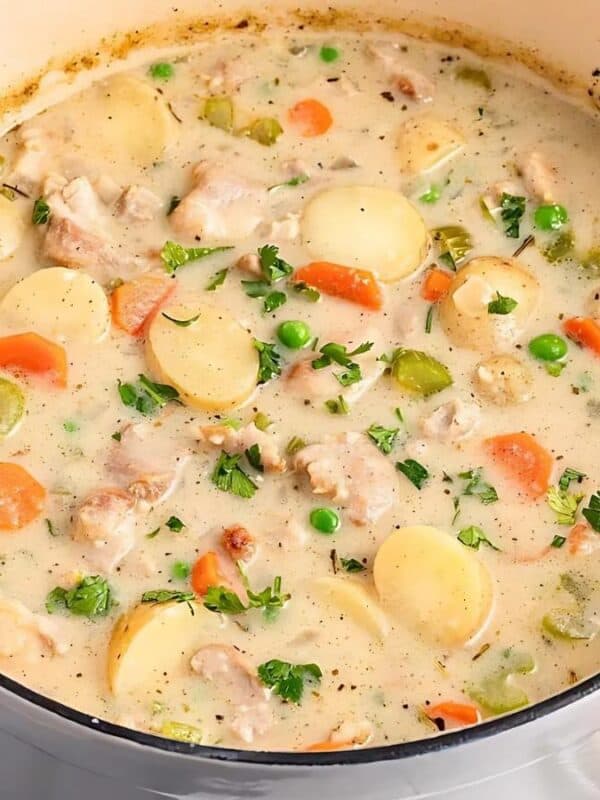 A creamy chicken stew in a white pot, filled with sliced carrots, peas, potatoes, and celery. The stew is garnished with chopped herbs. In the background are salt and pepper shakers and sprigs of fresh herbs. A slice of bread is on the side.