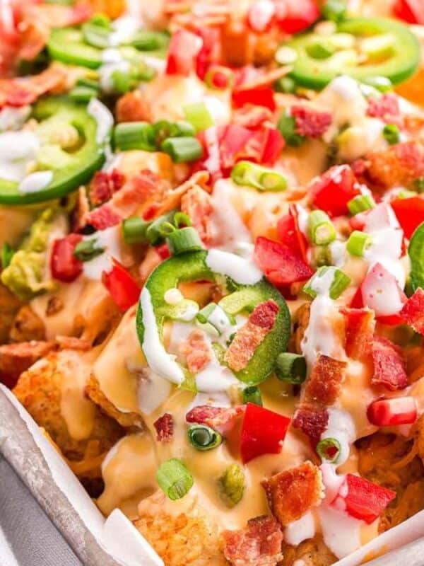 A tray of loaded tater tots topped with sliced jalapeños, diced tomatoes, green onions, crispy bacon bits, and drizzled with melted cheese and sour cream. A green and white checkered napkin is beside the tray on a wooden surface.