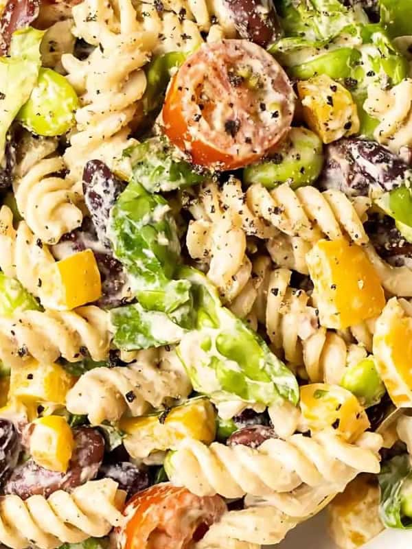 White bowl of pasta salad. The pasta is rotini, and the salad also contains kidney beans, edamame, avocado, cherry tomatoes, and a creamy dressing. Two gold forks are in the bowl. The background is a light gray surface. A white and gray striped napkin is partially visible in the lower left corner. A tomato is partially visible in the upper left corner.
