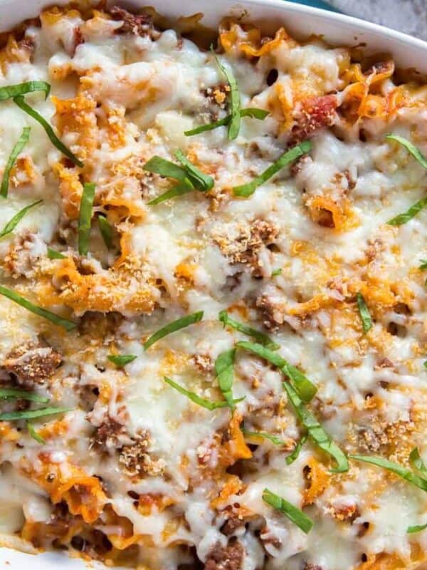 A baked pasta dish in a white oval dish, topped with melted cheese and garnished with chopped fresh herbs. The edges of the pasta are slightly browned, and it's placed on a blue cloth with a cutting board nearby.