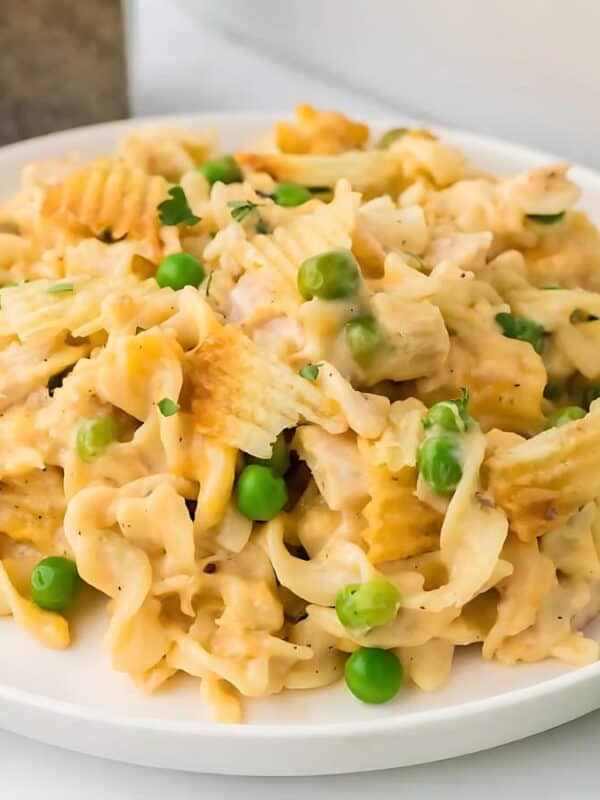 A creamy pasta dish with rotini noodles, green peas, and ruffled potato chips on top, served on a white plate. Garnished with fresh herbs.