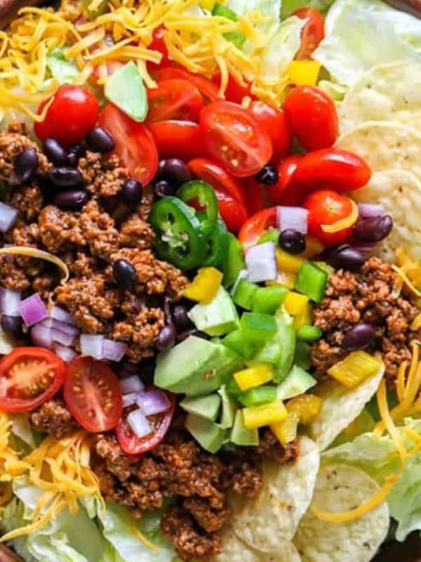 A colorful taco salad in a wooden bowl, topped with ground meat, cherry tomatoes, black beans, jalapeños, diced onions, yellow peppers, shredded cheese, and tortilla chips. Sliced avocado and salsa are on the side.