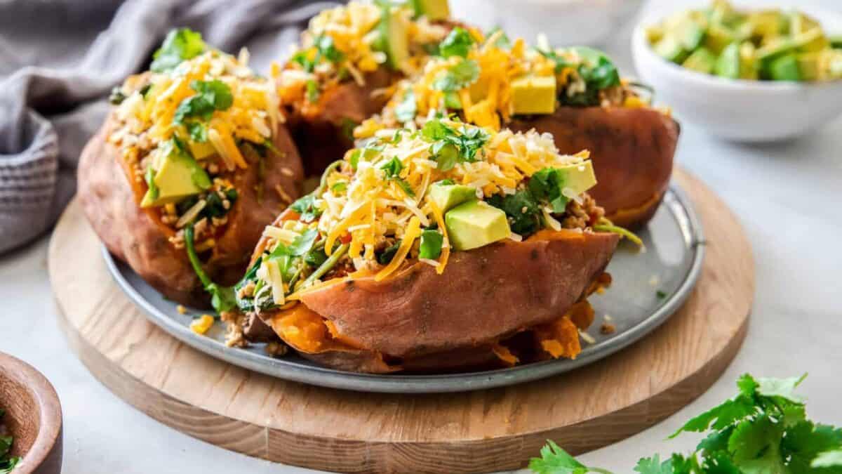 A plate of baked sweet potatoes topped with diced avocado, shredded cheese, and fresh cilantro. In the background, a small bowl contains more toppings. The dish is set on a wooden board with a soft cloth nearby.