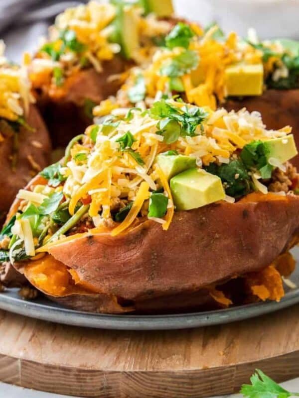 A plate of baked sweet potatoes topped with diced avocado, shredded cheese, and fresh cilantro. In the background, a small bowl contains more toppings. The dish is set on a wooden board with a soft cloth nearby.