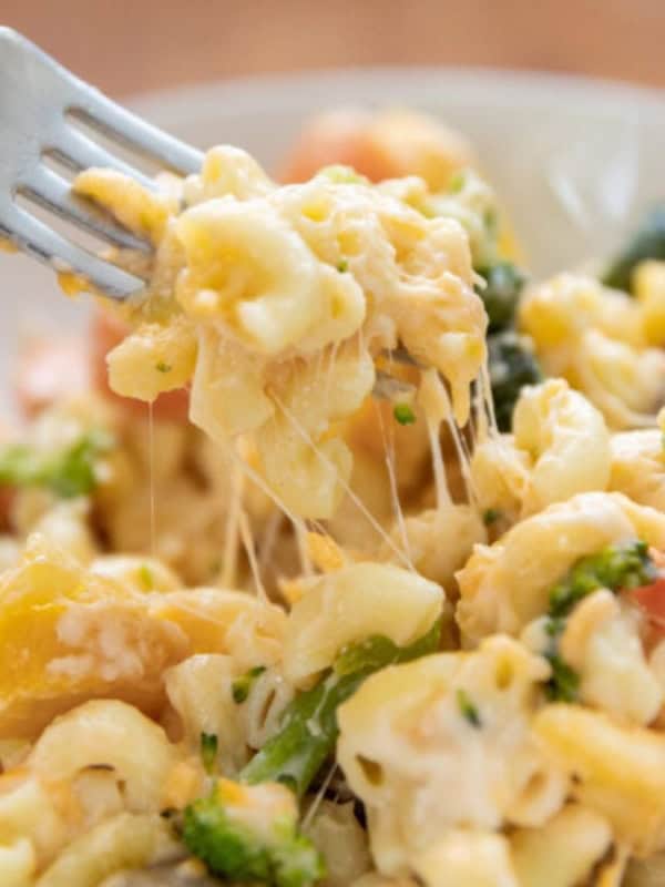 A fork lifts a cheesy serving of macaroni and vegetables from a plate. The pasta is mixed with broccoli, carrots, and yellow peppers, highlighting its creamy texture and colorful ingredients on a wooden table background.