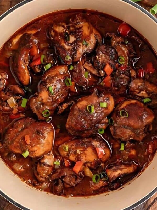A large pot filled with cooked chicken pieces in a rich, brown sauce garnished with chopped green onions. A bowl of rice and beans sits nearby, along with a wooden spoon and a cutting board with a red bell pepper.