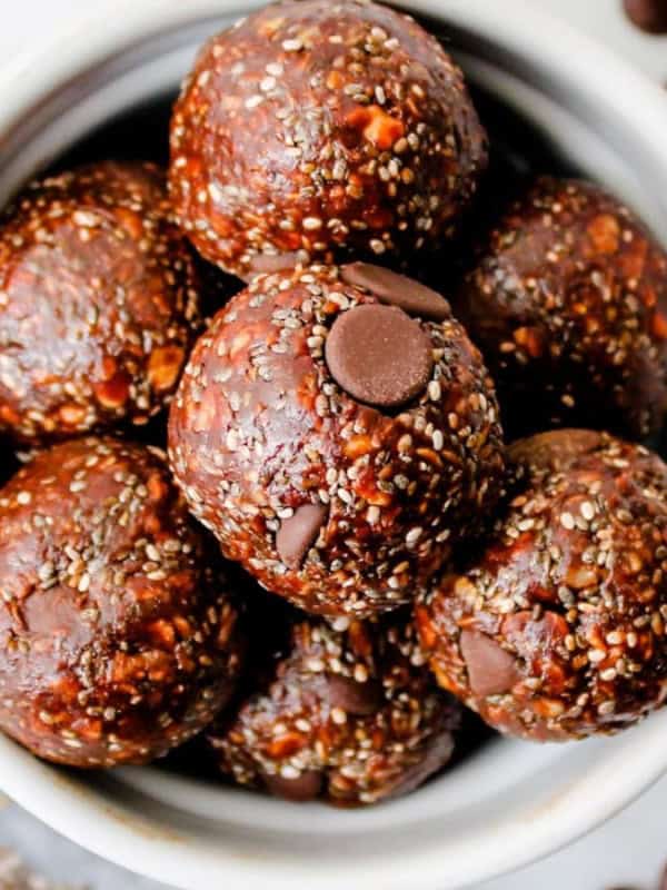 A top view of a bowl filled with chocolate energy balls, topped with chocolate chips and chia seeds. Around the bowl are scattered chia seeds and chocolate chips on a marble surface.