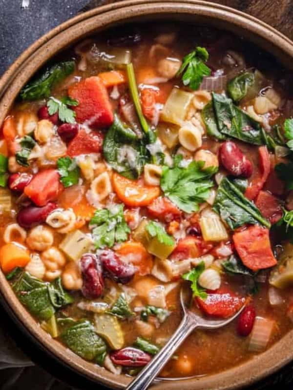A bowl of hearty vegetable soup with pasta, beans, and leafy greens, garnished with fresh parsley. A spoon rests inside the bowl, set on a dark surface with a light cloth.