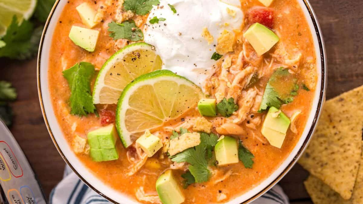 A bowl of creamy chicken tortilla soup topped with sliced limes, avocado cubes, cilantro, and a dollop of sour cream. The soup is a rich orange color, garnished with ground pepper, and there are tortilla chips on the side.