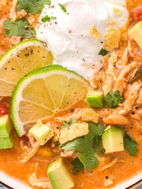 A bowl of creamy chicken tortilla soup topped with sliced limes, avocado cubes, cilantro, and a dollop of sour cream. The soup is a rich orange color, garnished with ground pepper, and there are tortilla chips on the side.