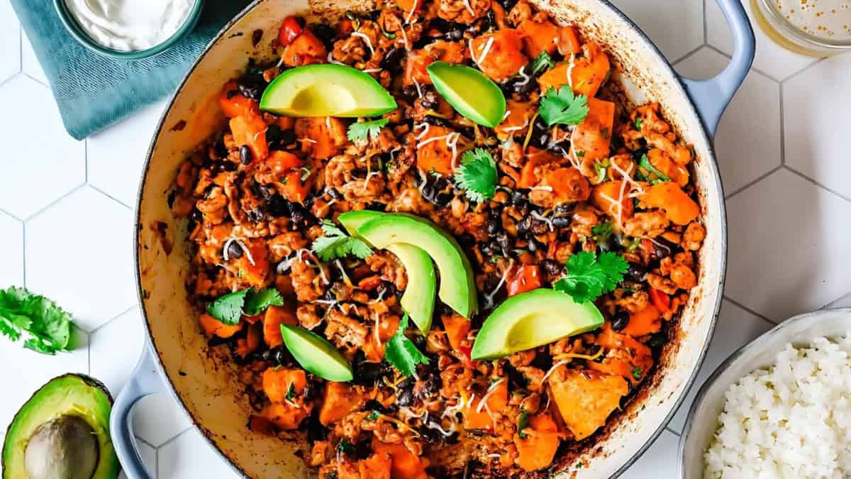 A skillet filled with a colorful dish featuring sweet potatoes, black beans, and ground meat, garnished with sliced avocado and cilantro. A small bowl of sour cream and a side of white rice are visible nearby on a hexagonal patterned table.