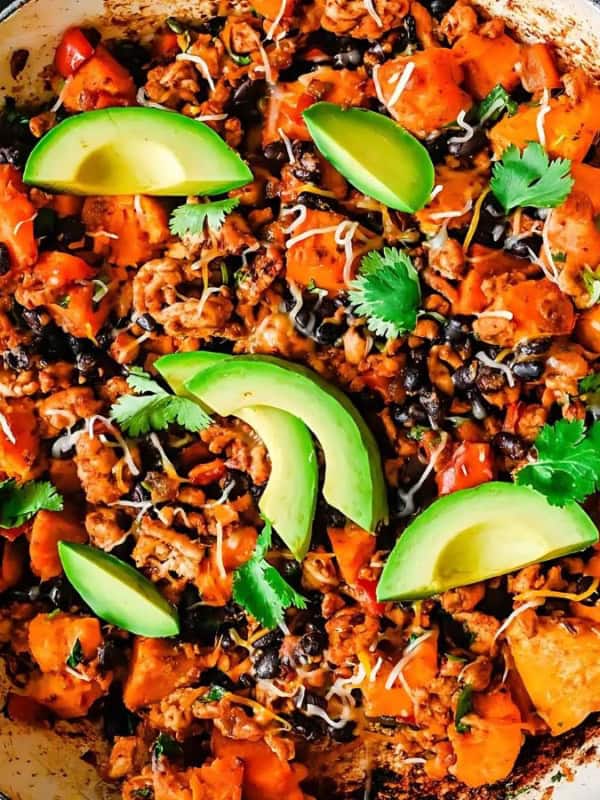 A skillet filled with a colorful dish featuring sweet potatoes, black beans, and ground meat, garnished with sliced avocado and cilantro. A small bowl of sour cream and a side of white rice are visible nearby on a hexagonal patterned table.