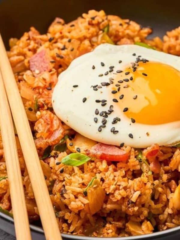 A bowl of fried rice topped with a sunny-side-up egg, sprinkled with black sesame seeds. Chopsticks rest on the side of the black bowl. The dish includes vegetables and appears garnished with green onions. A jar of sesame seeds is nearby.