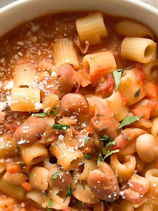 A bowl of pasta e fagioli featuring small pasta, beans, and vegetables in a rich tomato broth. Garnished with grated cheese and herbs, the dish is served in a white ceramic bowl on a white marble surface.
