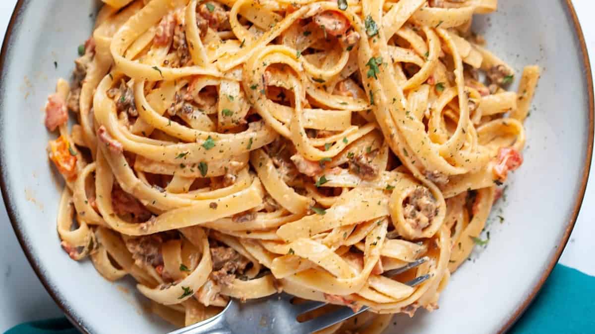 A plate of creamy pasta with a rich sauce, featuring fettuccine noodles mixed with minced meat, diced tomatoes, and garnished with herbs. A fork is twirled into the pasta, ready to be enjoyed.