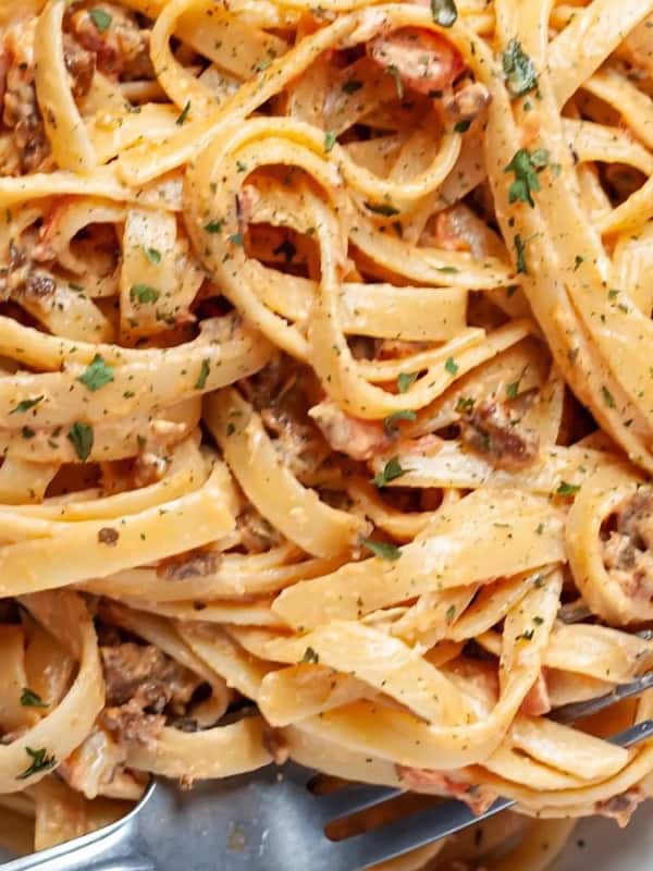 A plate of creamy pasta with a rich sauce, featuring fettuccine noodles mixed with minced meat, diced tomatoes, and garnished with herbs. A fork is twirled into the pasta, ready to be enjoyed.