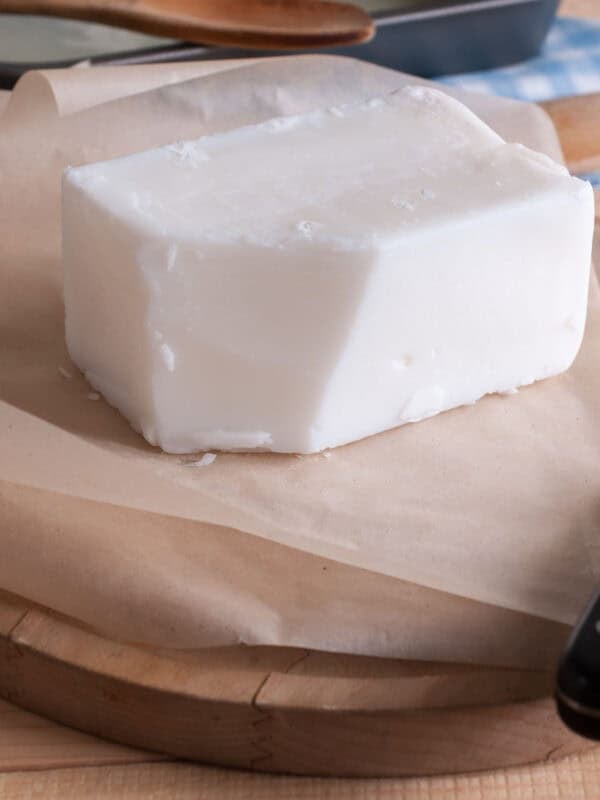 Solid block of white shortening on brown parchment paper placed on a round wooden board. A black-handled knife and a wooden spoon are partially visible in the background.