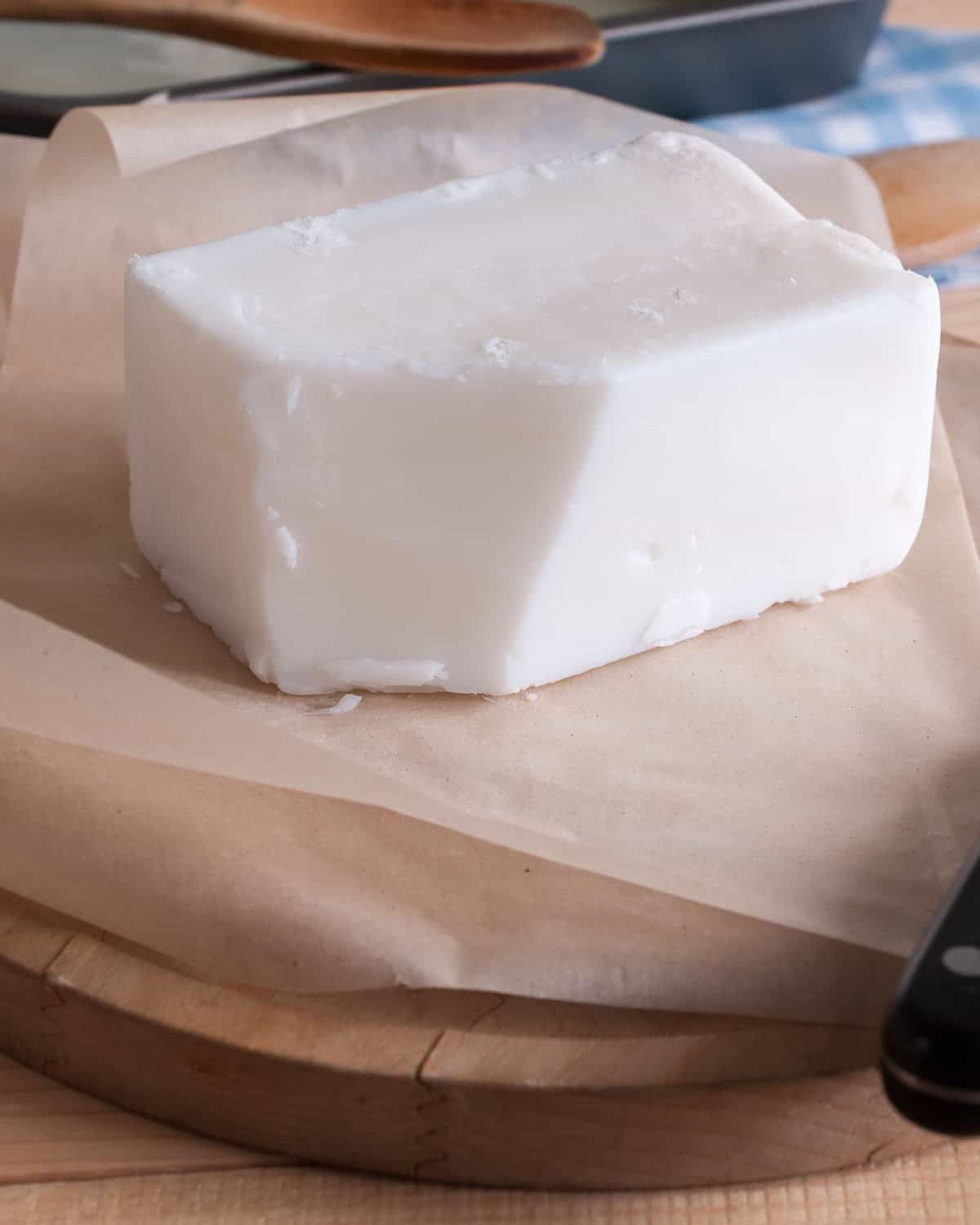 Solid block of white shortening on brown parchment paper placed on a round wooden board. A black-handled knife and a wooden spoon are partially visible in the background.