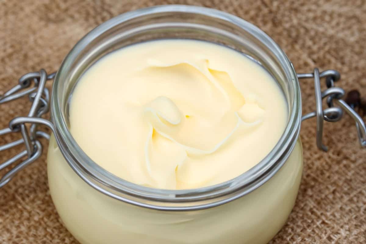 A glass jar filled with smooth, creamy white mayonnaise is placed on a textured burlap surface. The mayonnaise appears thick and glossy, with swirls on the surface. The jar has a metal clasp and a rubber seal.