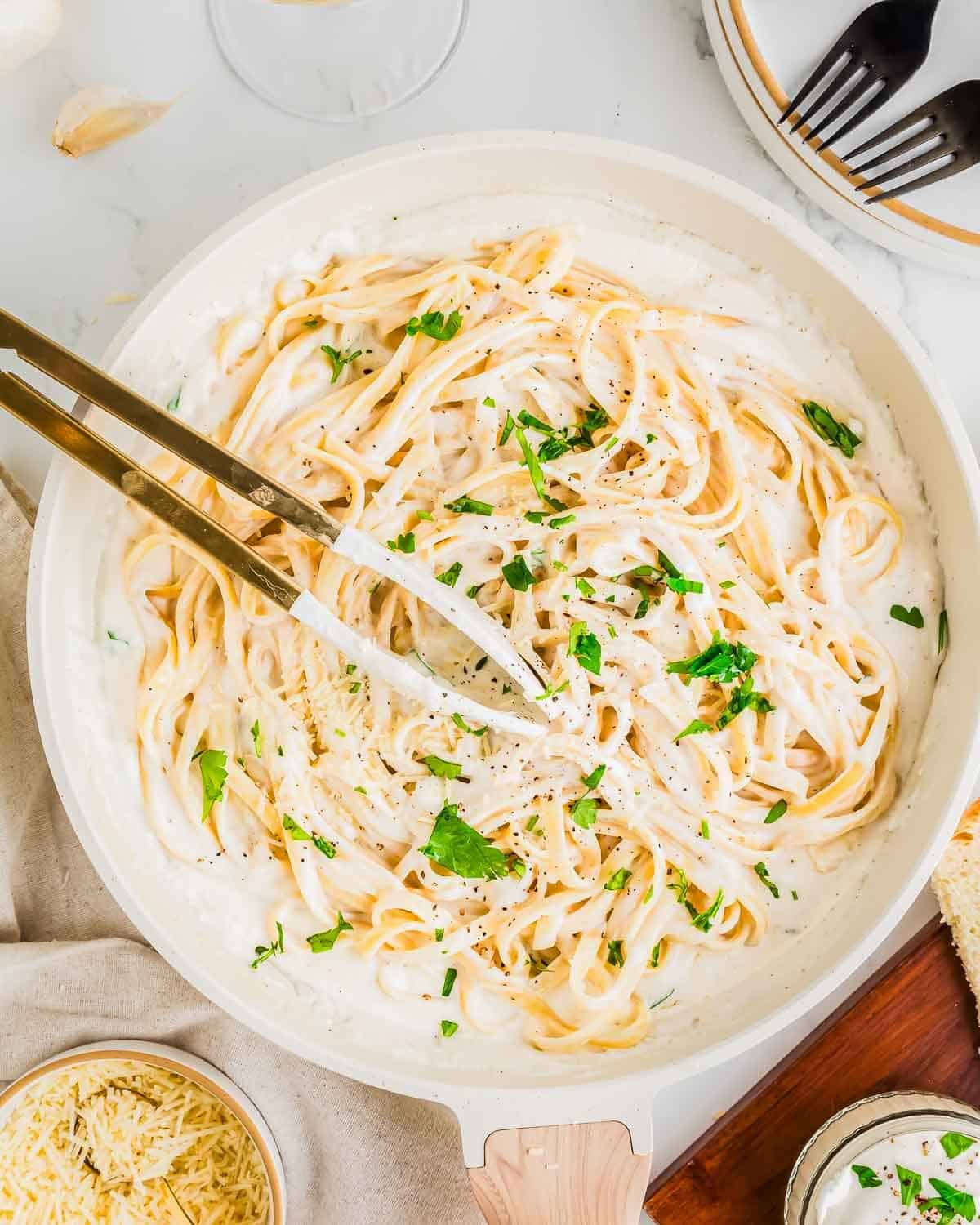 A white pan filled with creamy cottage cheese Alfredo fettuccine topped with chopped parsley and freshly ground black pepper. A pair of tongs rests in the pan. Nearby, a small bowl of grated cheese and utensils are visible on a light-colored surface.