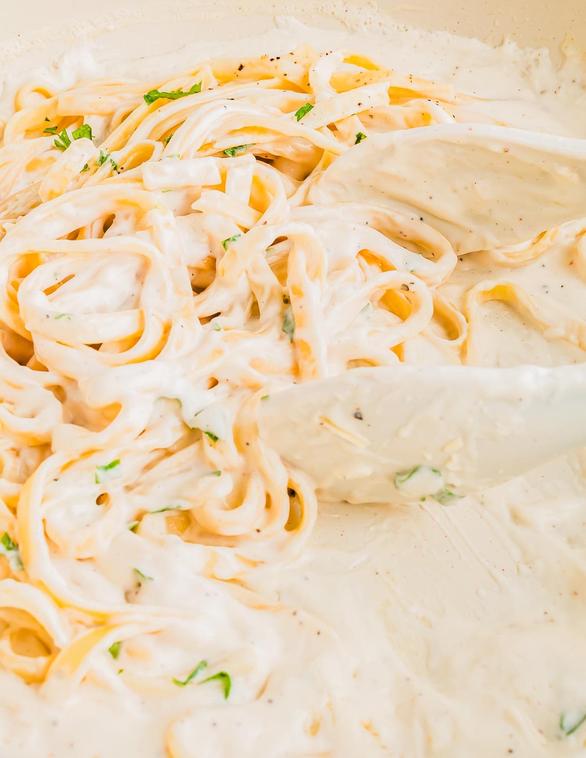 Close-up of creamy pasta being stirred with a spatula. The noodles are coated in a thick, Alfredo sauce with visible flecks of herbs, giving it a rich and indulgent appearance.