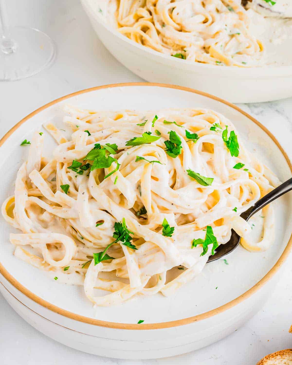 A plate of fettuccine cottage cheese Alfredo garnished with chopped parsley. The creamy pasta is served on a white plate with a fork. A bowl of additional pasta is visible in the background.