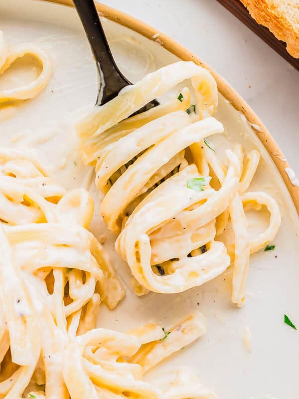 A plate of creamy pasta with a fork twirling the noodles; garnished with herbs. Two slices of bread are placed beside the plate on a wooden board.