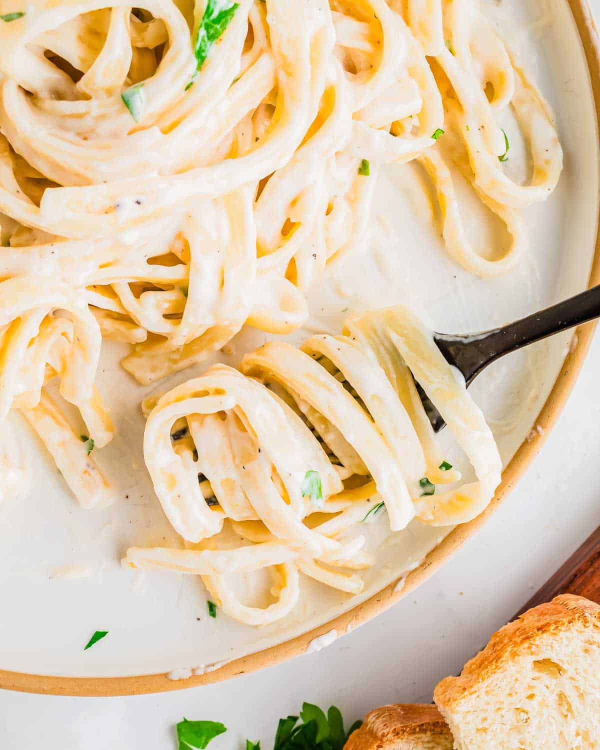 A close-up of creamy fettuccine Alfredo pasta garnished with parsley on a plate. A fork holds a twirl of pasta. A slice of toasted bread is partially visible on the side of the dish.