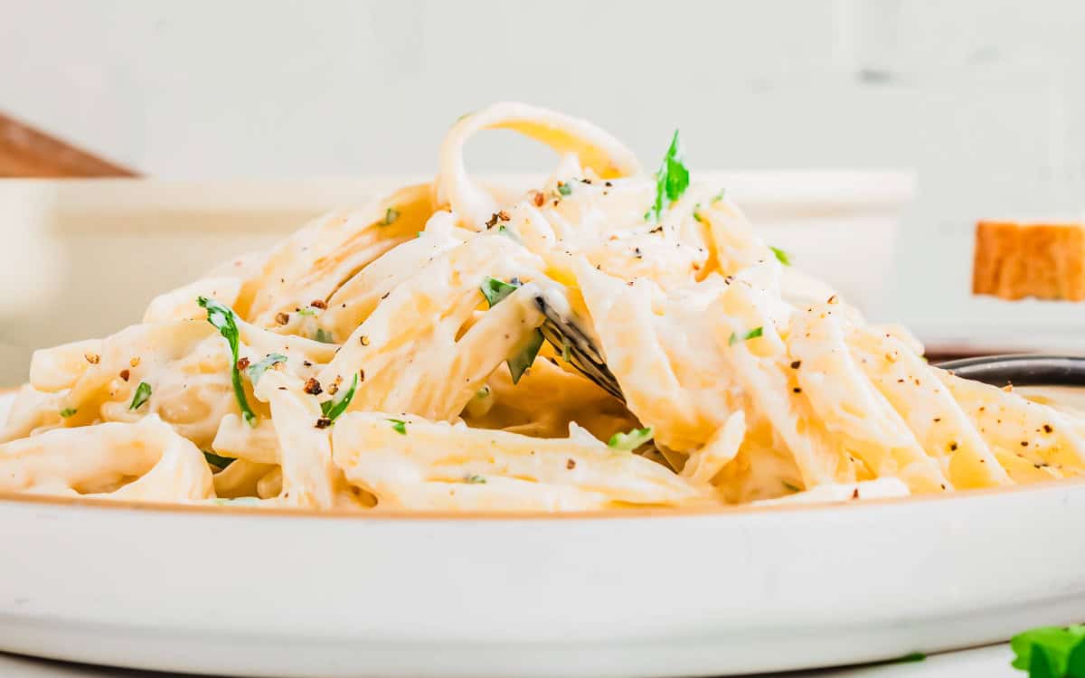 A close-up of creamy fettuccine cottage cheese Alfredo on a white plate, garnished with parsley and black pepper. A fork is twirling some pasta, with a blurred background adding depth to the image.
