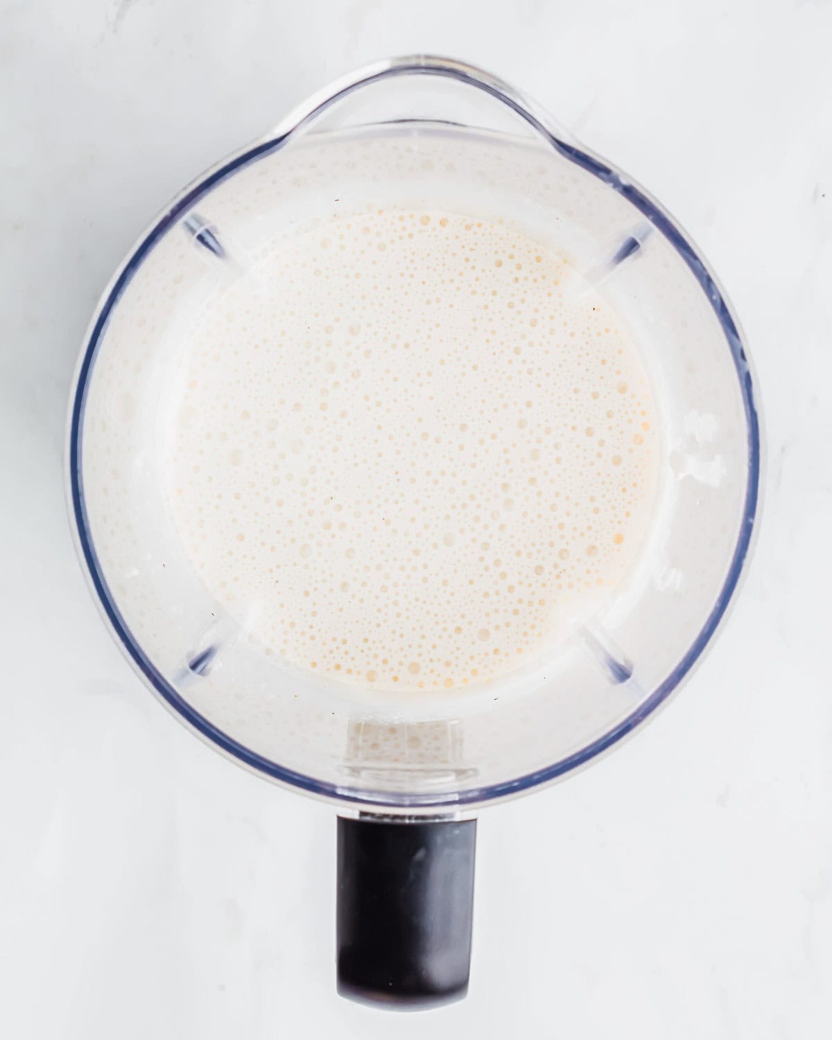 Top view of a blender filled with a creamy cottage cheese sauce on a white surface. The blender has a black handle, and the liquid appears smooth and evenly mixed, suggesting it could be a smoothie or a similar blended drink.