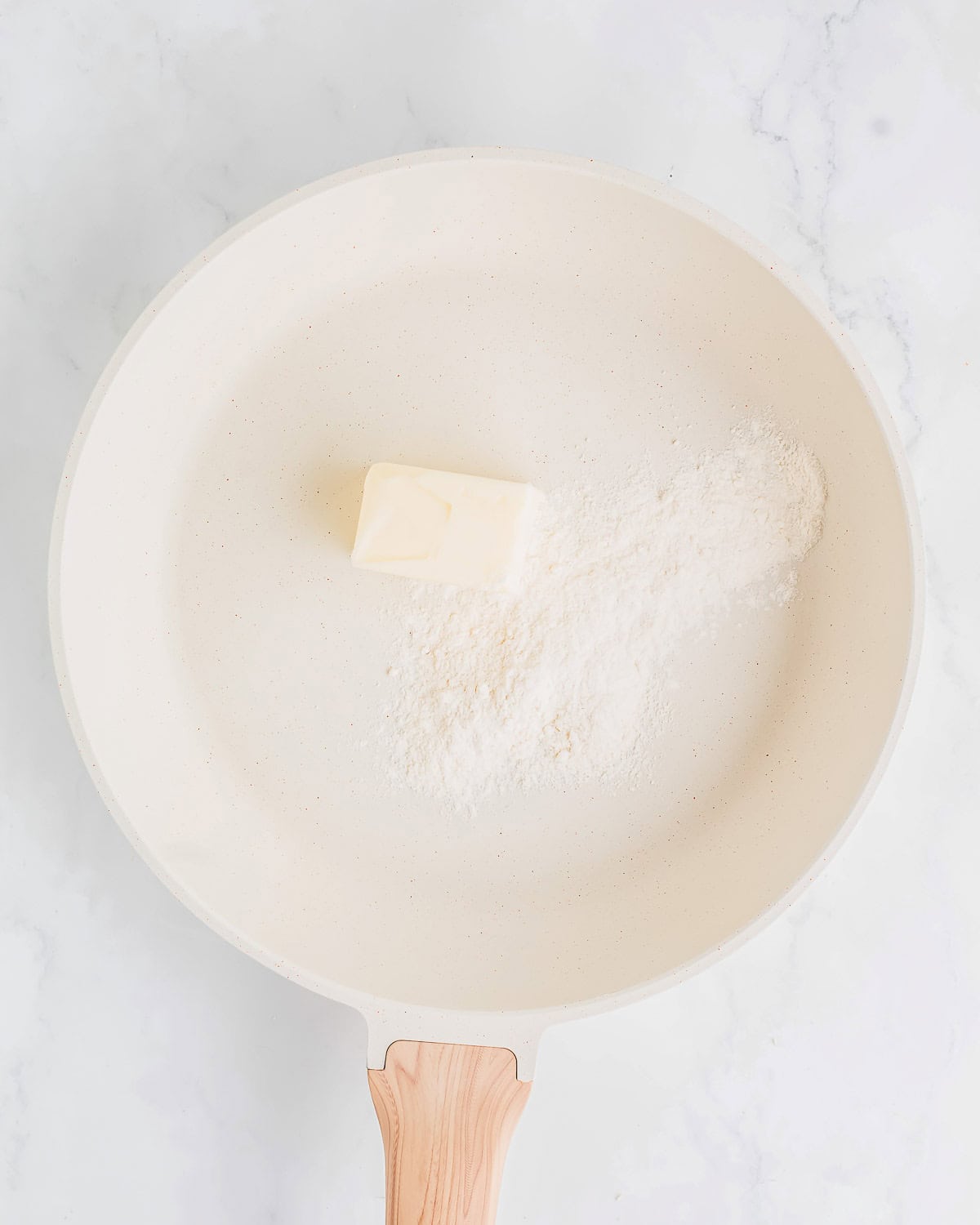 A white frying pan on a marble surface contains a small pile of flour and a pat of butter, ready for cooking. The pan has a wooden handle.