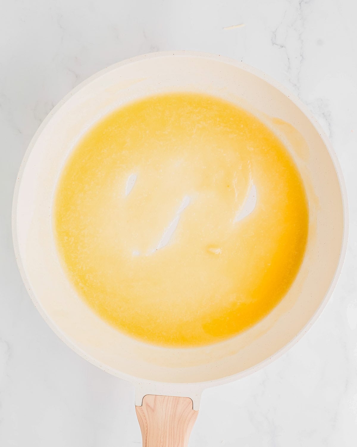 A top view of a frying pan with melted butter forming a yellow pool. The pan has a light-colored handle and is placed on a white marble surface. Bright lighting highlights the smooth, glossy texture of the butter.