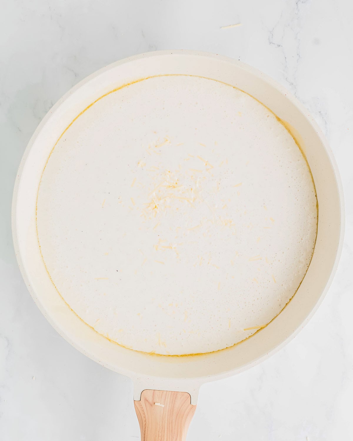 A white pan with a wooden handle is filled with a creamy white cottage cheese sauce, topped with a small sprinkle of grated cheese, against a marble surface.