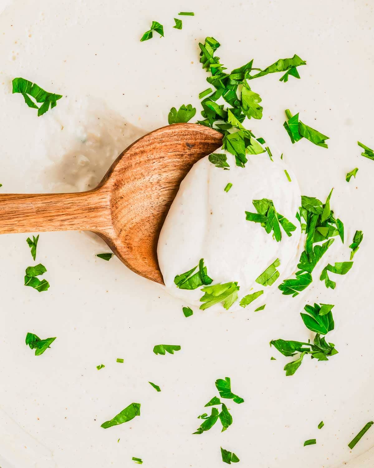 A wooden spoon holds a dollop of creamy Alfredo sauce made with cottage cheese, garnished with fresh, chopped green parsley. The sauce is spread on a flat surface, with parsley scattered around.