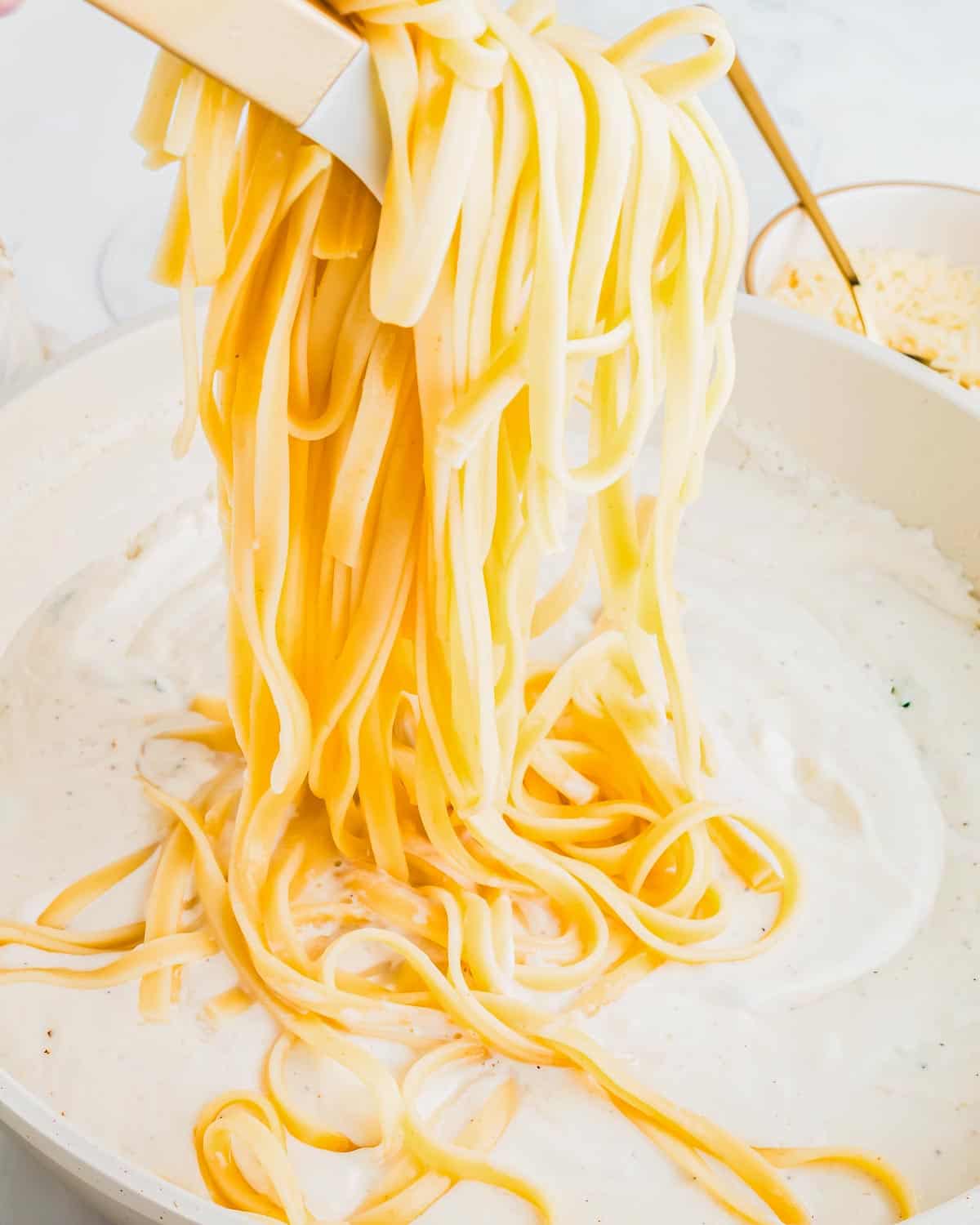 Pasta being lifted from a pot filled with Alfredo sauce. The scene captures the silky texture of the pasta and the richness of the sauce. In the background, there's a glimpse of a bowl with grated cheese.