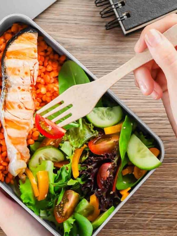 A person uses a wooden fork to eat from a meal prep container containing grilled salmon, lentils, and a colorful salad with tomatoes, cucumbers, and greens. Nearby are a laptop, notebook, container with eggs, and an apple.