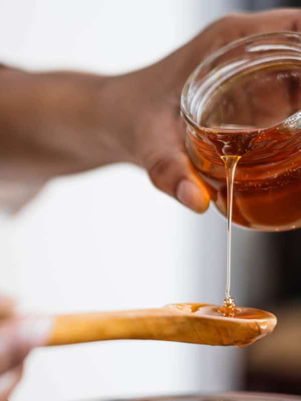 A person holding a jar of honey, pouring it onto a wooden spoon. The focus is on the honey as it flows from the jar to the spoon, with a blurred background.