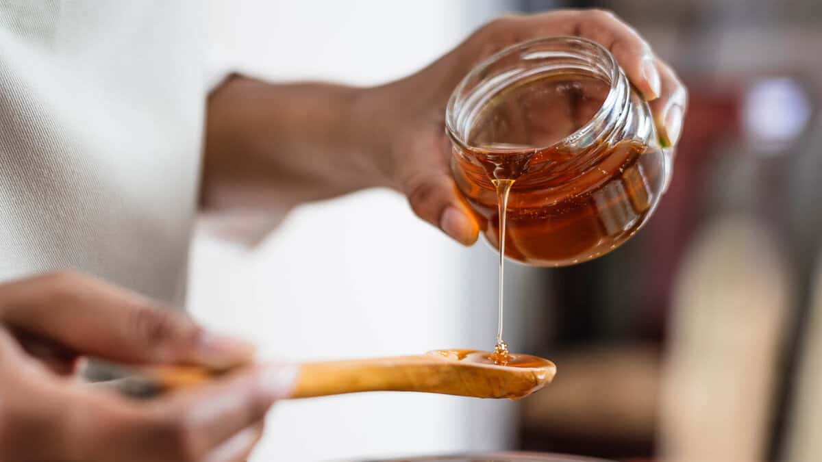A person holding a jar of honey, pouring it onto a wooden spoon. The focus is on the honey as it flows from the jar to the spoon, with a blurred background.