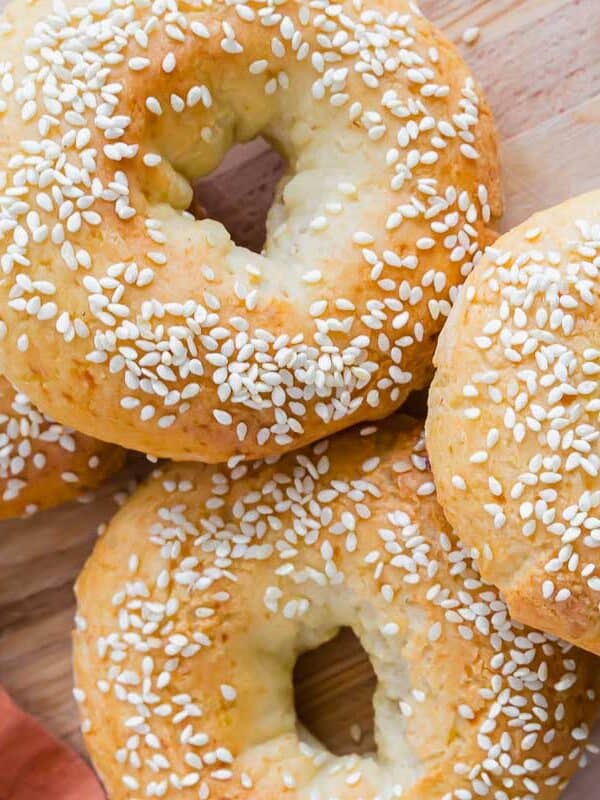 Four golden-brown bagels topped with sesame seeds are arranged on a wooden board. A piece of orange fabric is visible on the left side.