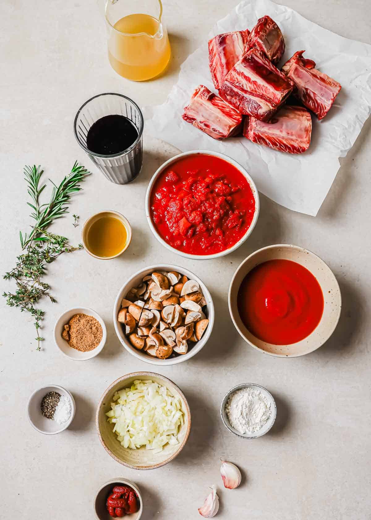 A flat lay of cooking ingredients including beef short ribs, crushed tomatoes, tomato paste, mushrooms, chopped onions, garlic, herbs, broth, olive oil, Worcestershire sauce, and seasoning arranged on a light surface.