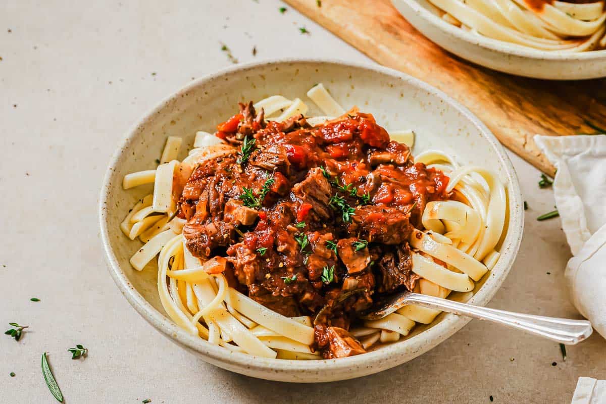 A bowl of tagliatelle pasta topped with a rich tomato and beef ragout, garnished with fresh herbs. A fork rests in the bowl, and the dish is set on a light-colored table. Another bowl of pasta can be seen in the background.