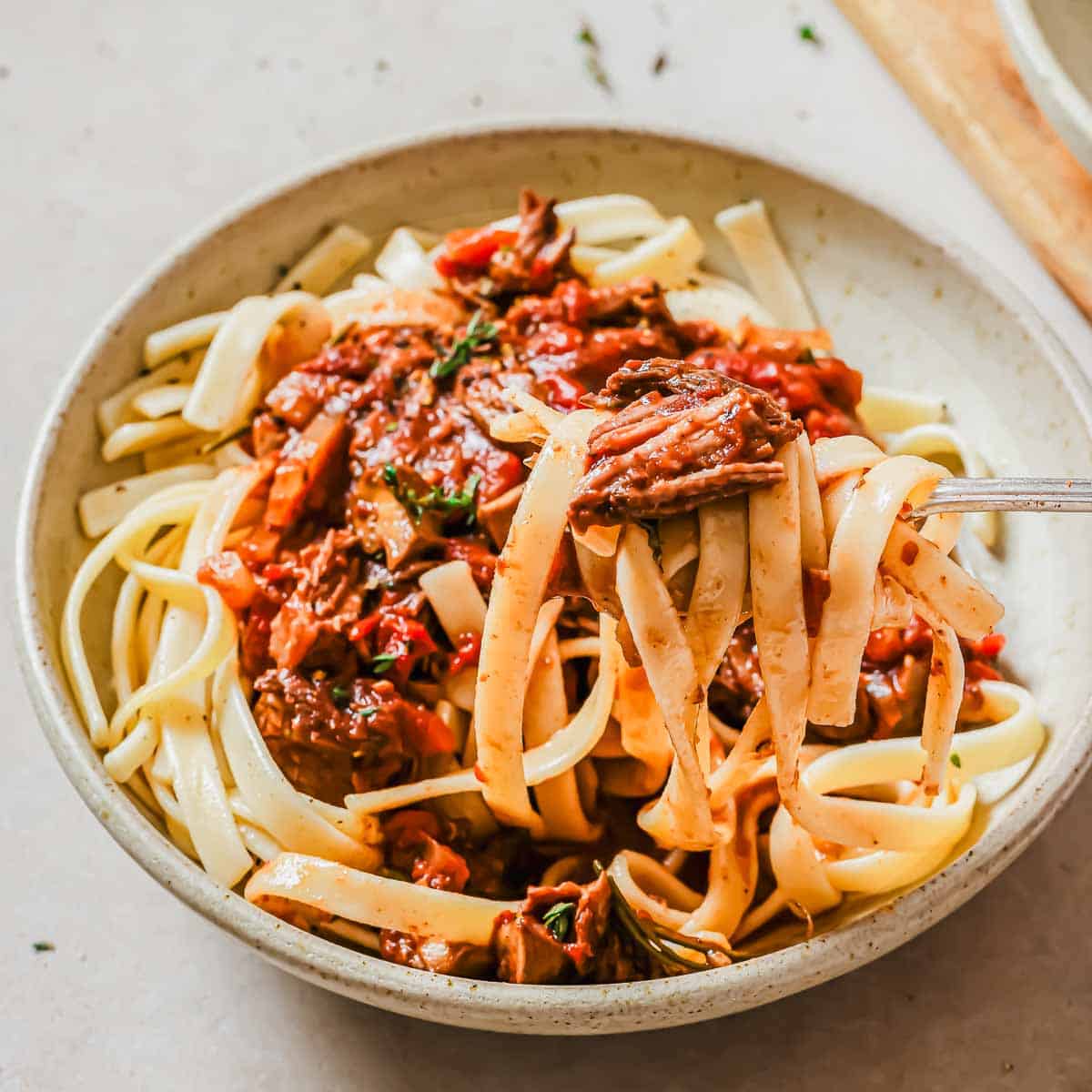 A bowl of tagliatelle pasta topped with a rich, savory short rib meat sauce with visible herbs and red pepper flakes. A fork is lifting a portion of pasta, showcasing the sauce and tender meat pieces. The dish is served in a neutral-toned bowl.