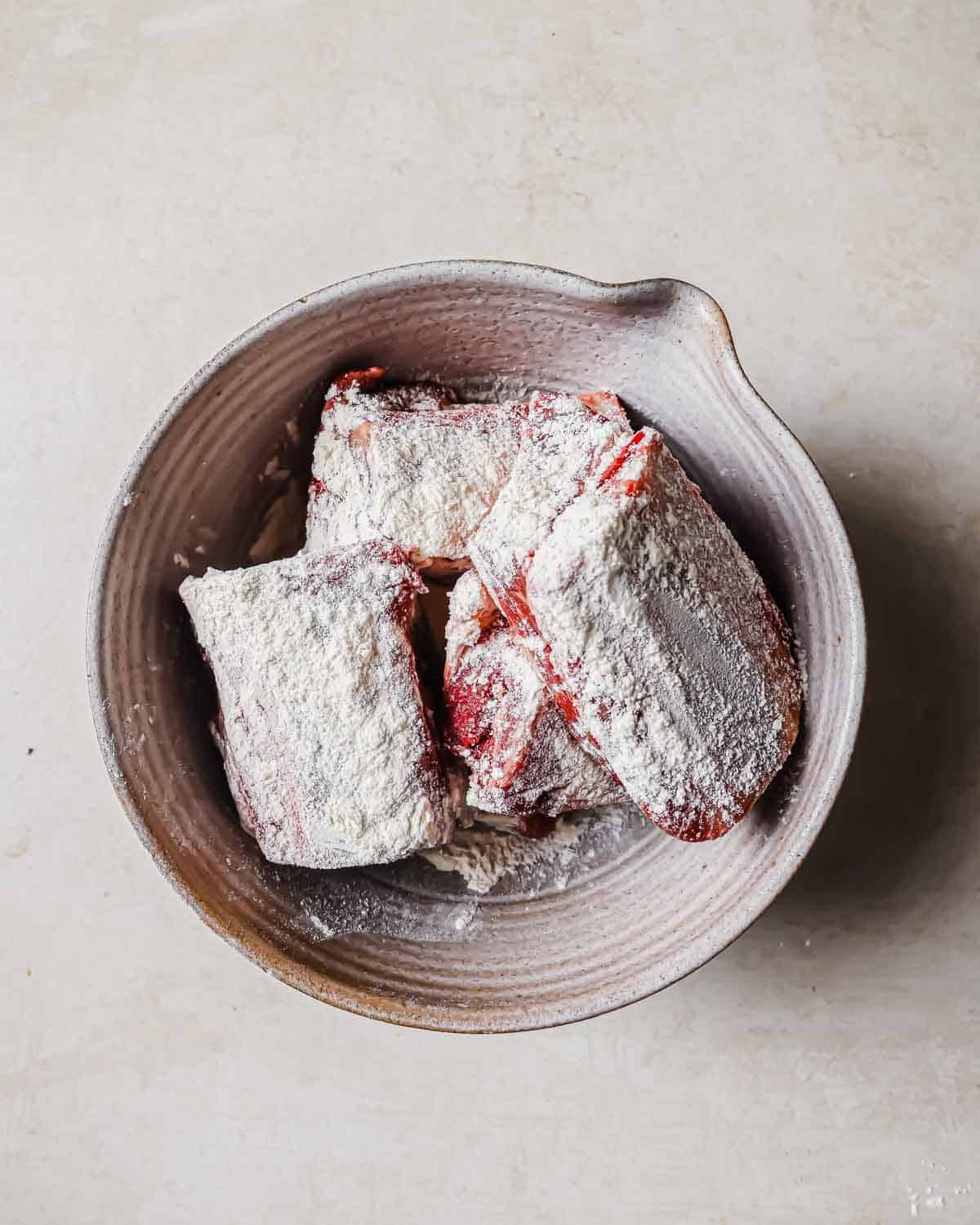 A bowl containing short rib pieces coated in flour, placed on a light-colored surface. The bowl has a rustic, textured appearance.