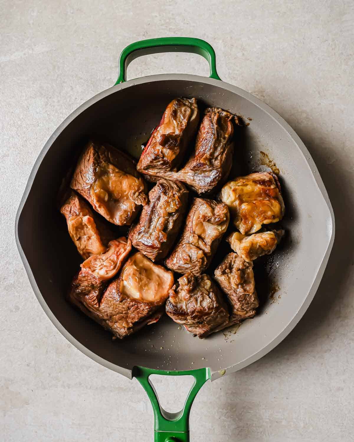 Browned beef short ribs in a green-handled skillet on a light surface. The ribs are browned and arranged neatly in the pan.