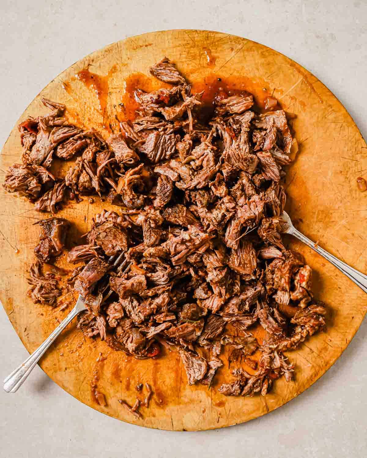 Shredded beef short ribs on a wooden cutting board with two forks, scattered in a rustic presentation. The meat appears juicy and tender, with rich brown hues, hinting at a savory dish ready to be served.