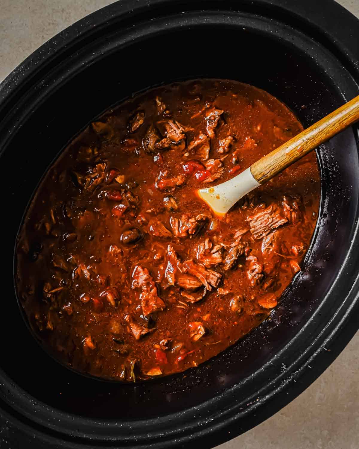 A slow cooker filled with a hearty short rib ragu. The stew is rich and red, featuring chunks of beef and vegetables, and is being stirred with a wooden spoon.