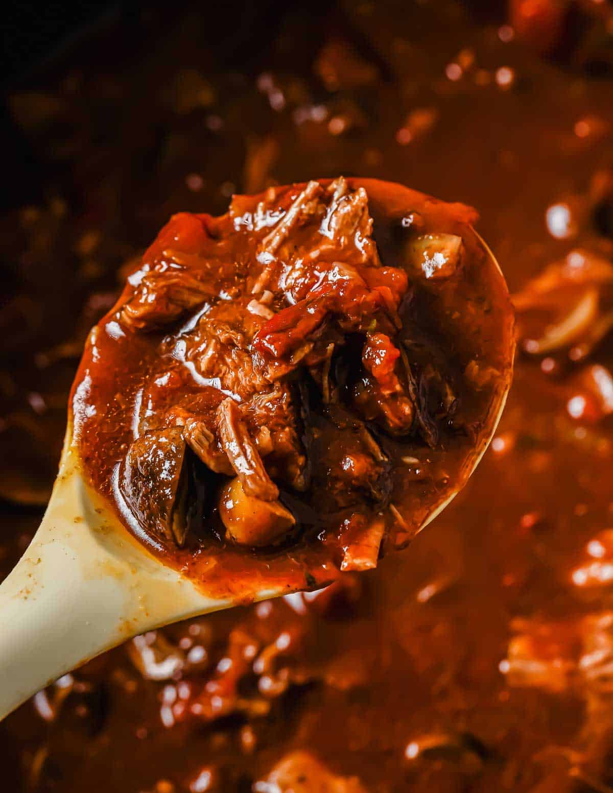 A close-up of a ladle filled with a rich, beef short rib ragu featuring chunks of beef, sliced onions, and tomatoes, all immersed in a thick, flavorful sauce. The ragu's surface glistens, hinting at its savory tenderness.
