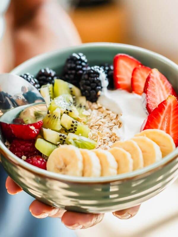 A person holds a bowl filled with yogurt, granola, sliced bananas, strawberries, kiwi, raspberries, and blackberries. A spoon is poised above the bowl, ready to take a bite. The background is softly blurred.