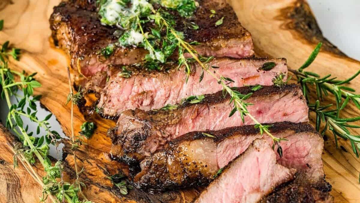 Sliced steak cooked to medium rare is garnished with herbs and a dollop of herb butter on a wooden serving board. Fresh sprigs of rosemary and thyme are arranged around the steak.