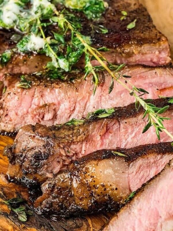 Sliced steak cooked to medium rare is garnished with herbs and a dollop of herb butter on a wooden serving board. Fresh sprigs of rosemary and thyme are arranged around the steak.